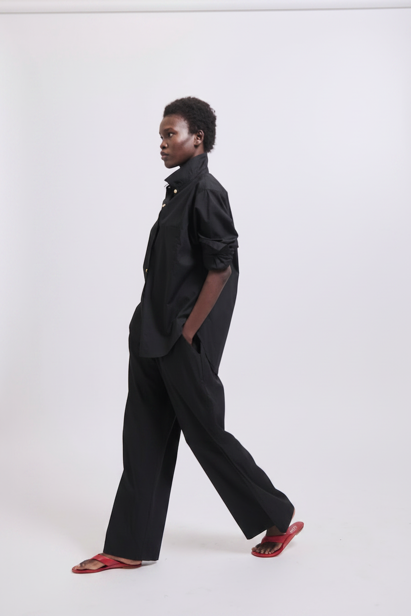 A Black woman in a black shirt and pants walks in red sandals against a white background.