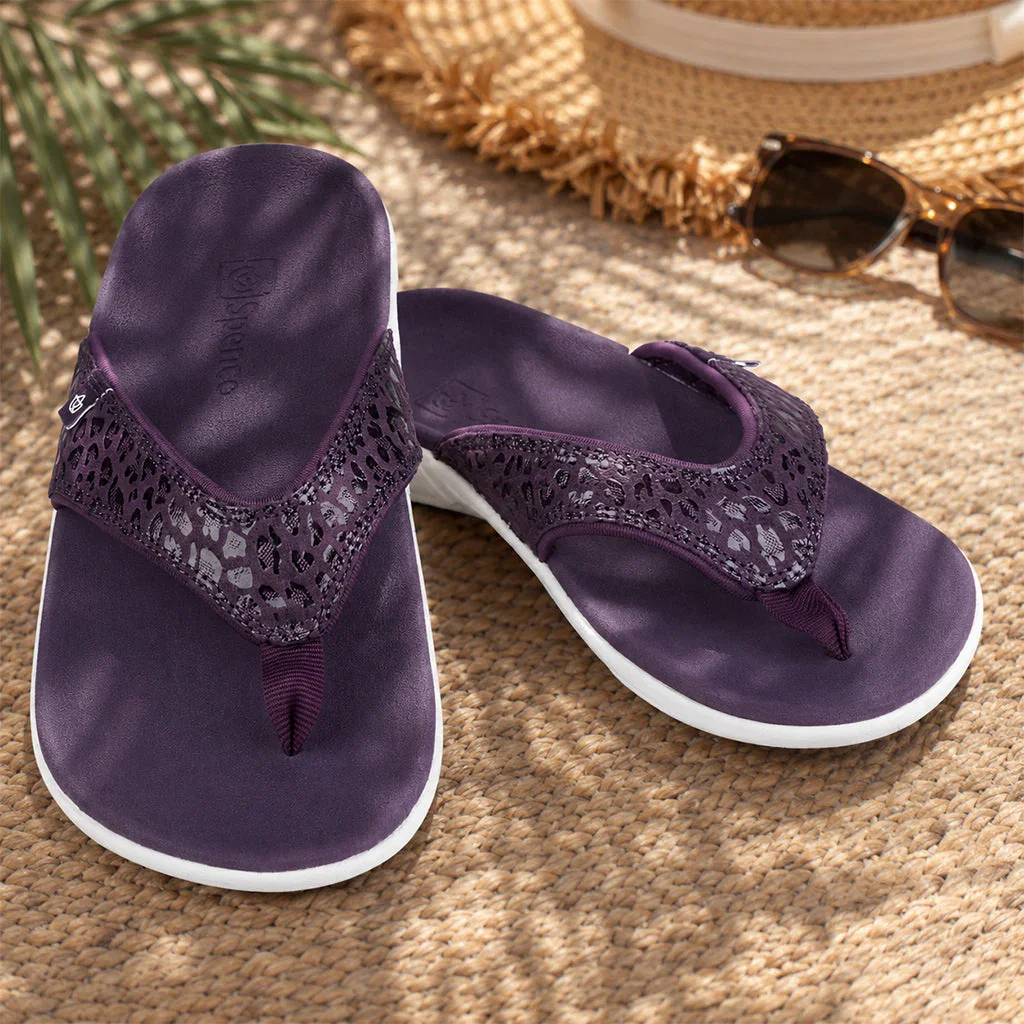 Pair of purple Spenco flip-flops with lace detailing on a beach setting with sunglasses and straw hat.