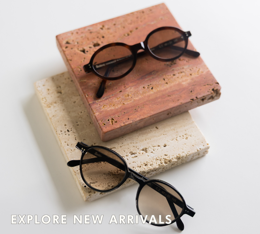 A stylized top-down product shot of two pairs of oval eyeglasses resting on stacked, square pieces of textured stone (one pink, one beige). The top pair is a dark tortoise-shell color, and the bottom pair is solid black. Transparent overlay text along the bottom reads: 