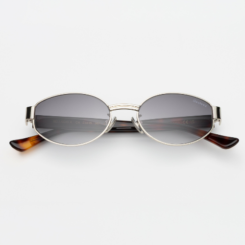 A studio product shot of the Bellagio sunglasses. These feature a shallow, oval gold metal frame with intricate textured detailing around the rims and bridge, paired with grey gradient lenses and tortoise-shell temple tips.