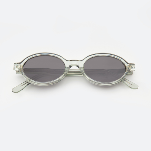 A studio product shot of oval-shaped sunglasses in a clear, translucent acetate with dark grey lenses.