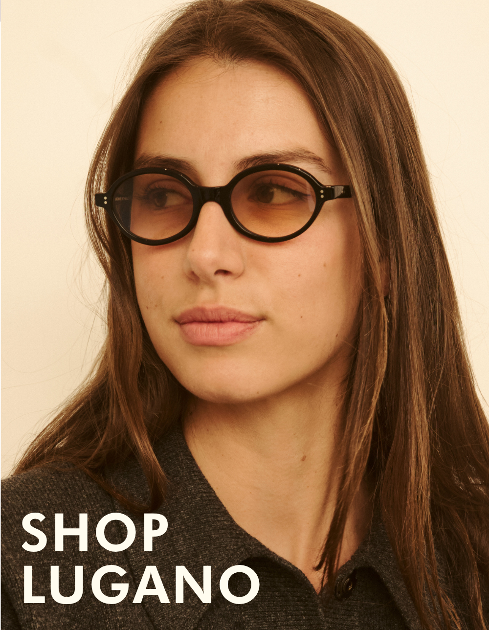 A close-up headshot of a woman with long brown hair looking off to the side. She is wearing the Lugano sunglasses, which feature a black oval frame with warm amber-to-purple gradient lenses. Bold white text in the corner reads: 