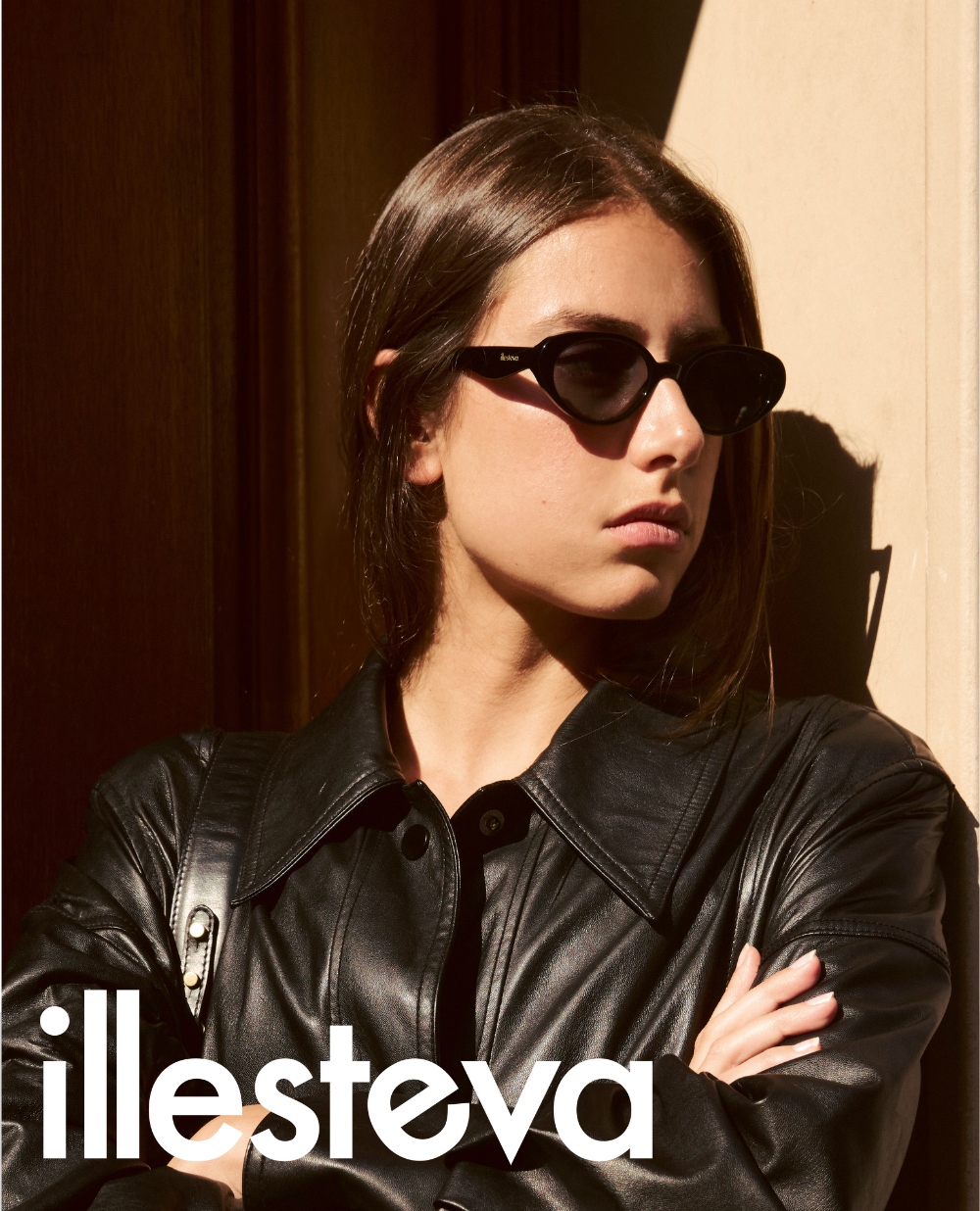 A campaign portrait of a woman leaning against a door frame in the sunlight. She is wearing a black leather jacket and small, pointed 90s-style black cat-eye sunglasses. The illesteva logo is overlayed in large white font at the bottom.
