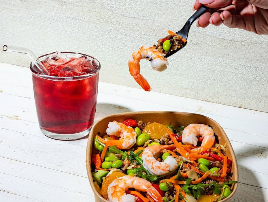 Photo of bowl with shrimp and vegetables, a drink with pink drink insite it, and a hand holding a fork 