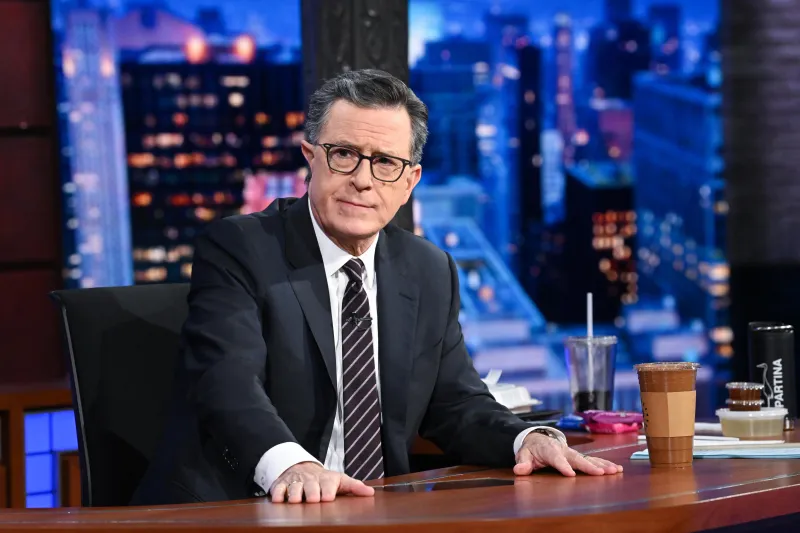 Stephen Colbert, with glasses, a black suit and striped tie, looking into the camera from his Late Show desk, with a to-go coffee at hand.