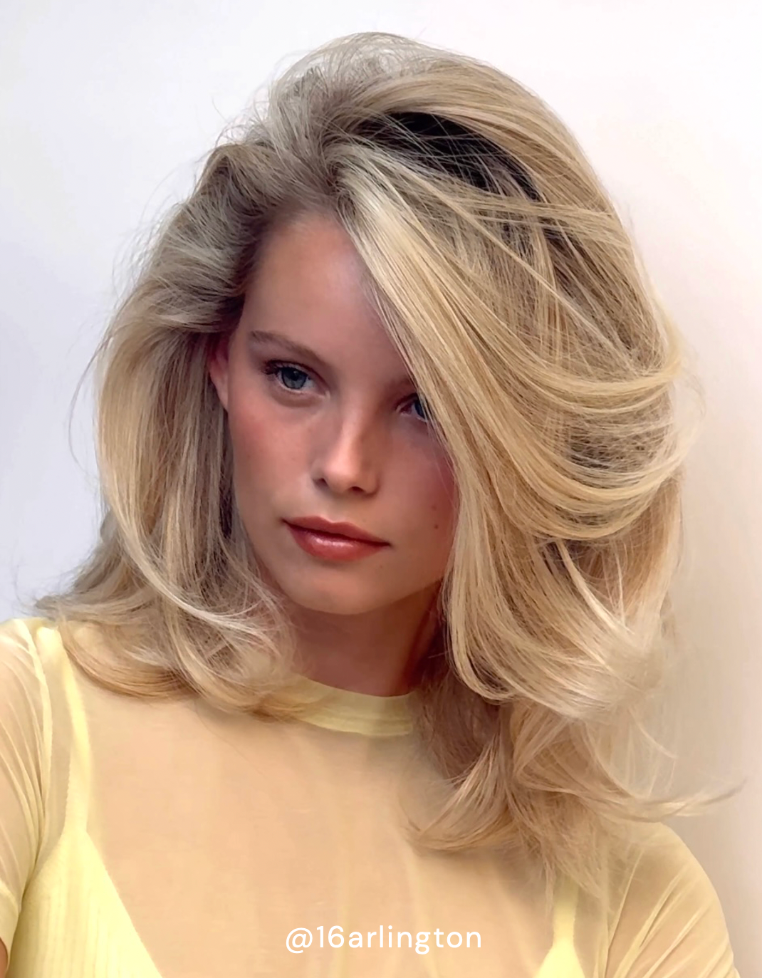 Woman with blonde hair styled in voluminous waves, wearing a sheer yellow top.