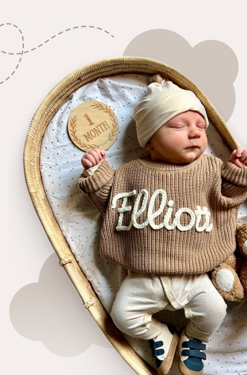 Newborn baby lying in a woven basket wearing a brown personalized sweater, white hat, and soft socks.