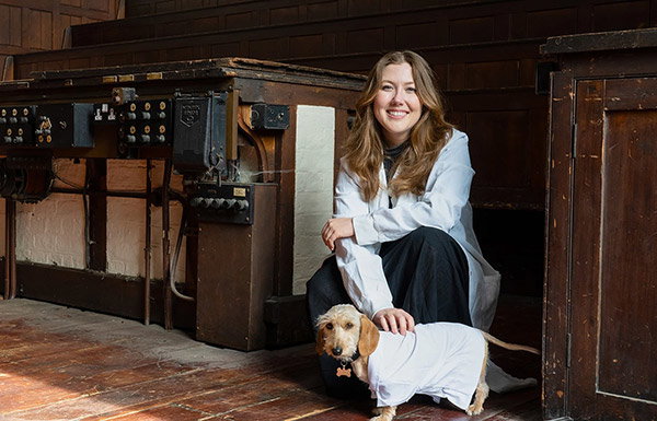 A scientist in a labcoat kneels next to a frankly adorable dog, also wearing a labcoat.