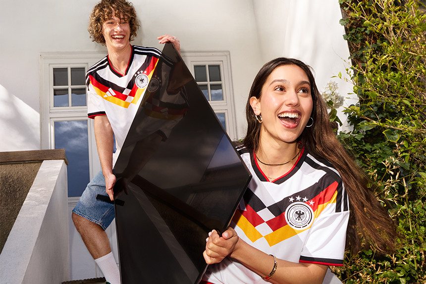 SaleZwei Personen in Deutschland-Trikots tragen gemeinsam einen großen Fernseher in den Garten.