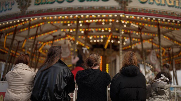 Four women stand in a row, with their backs to the camera, looking at a funfair carousel going round.