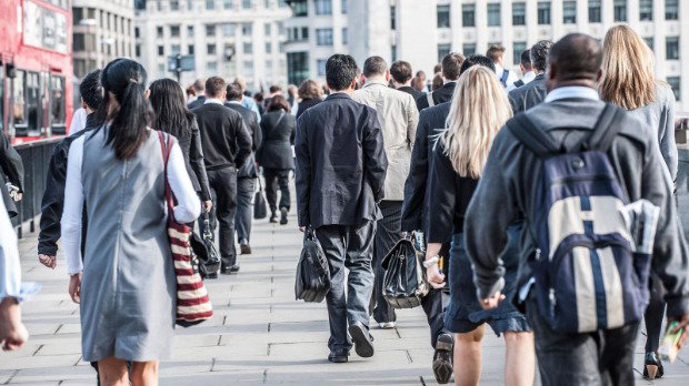 Lots of commuters walking along a pavement with their backs to the camera. Office buildings can be seen in the background.