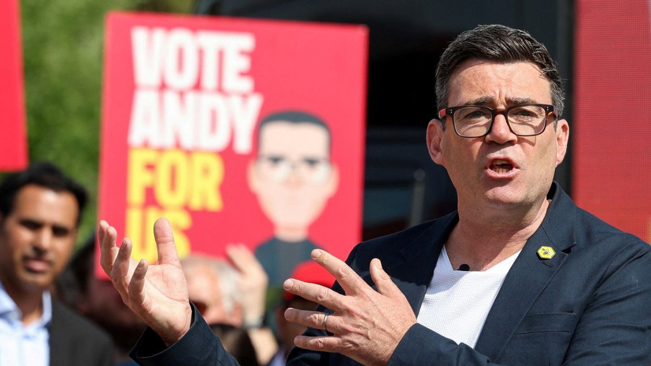 Andy Burnham speaks and gestures with his hands at a rally in Makerfield. He wears a white t shirt, jacket and glasses and has dark hair. Crowds and posters supporting him are behind him slightly blurred.