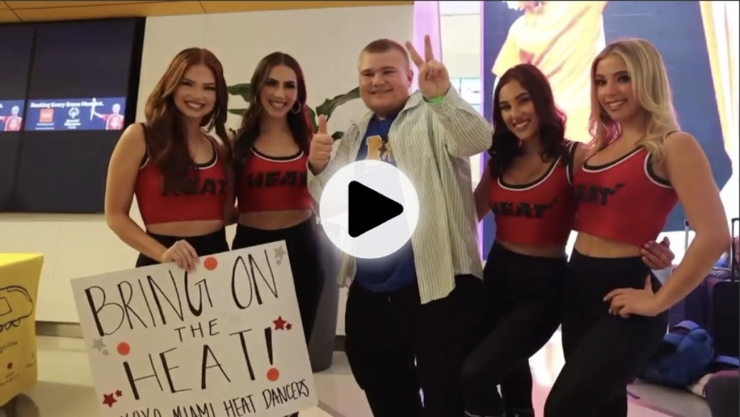 A man poses with a group of four cheerleaders wearing matching red crop tops and black pants. A large play button overlay suggests this is a video thumbnail.