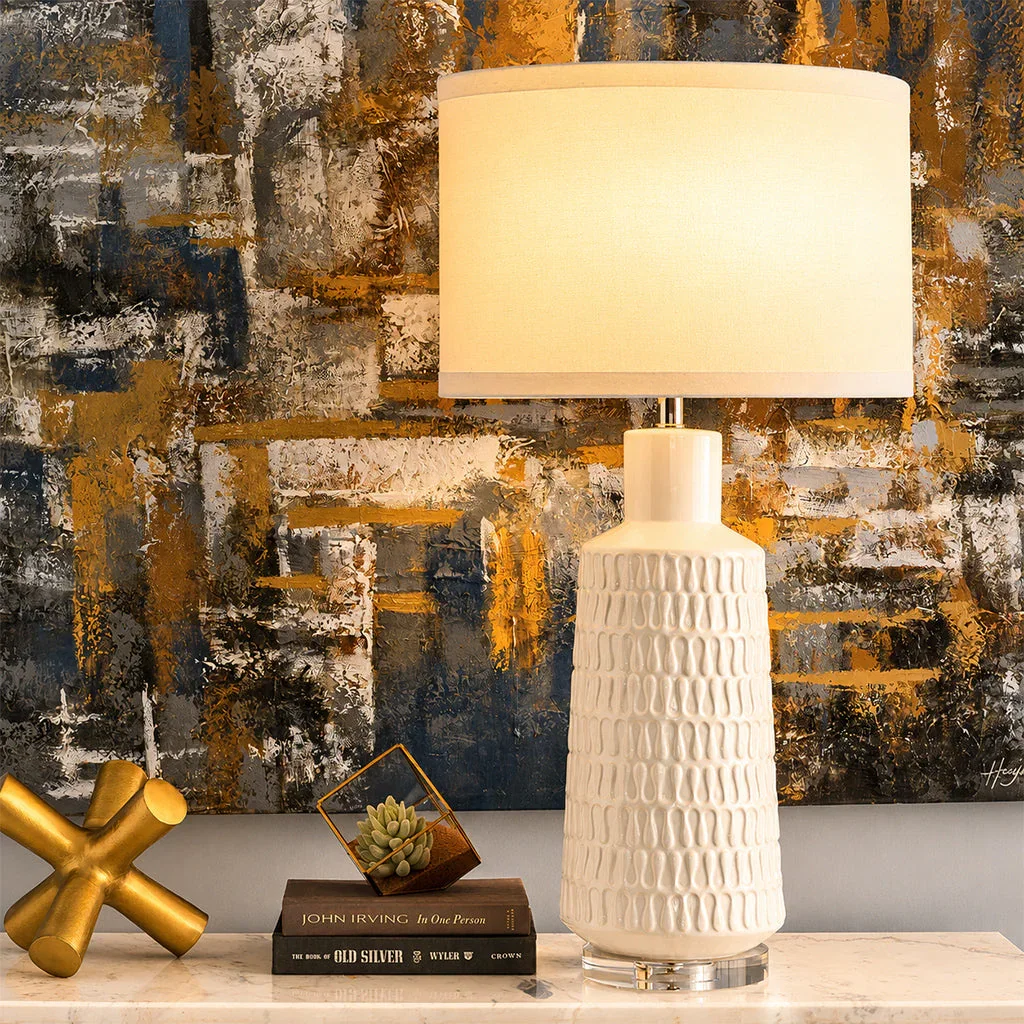 Elegant cream ceramic table lamp with textured base and beige shade, styled on marble surface with gold decor and books.