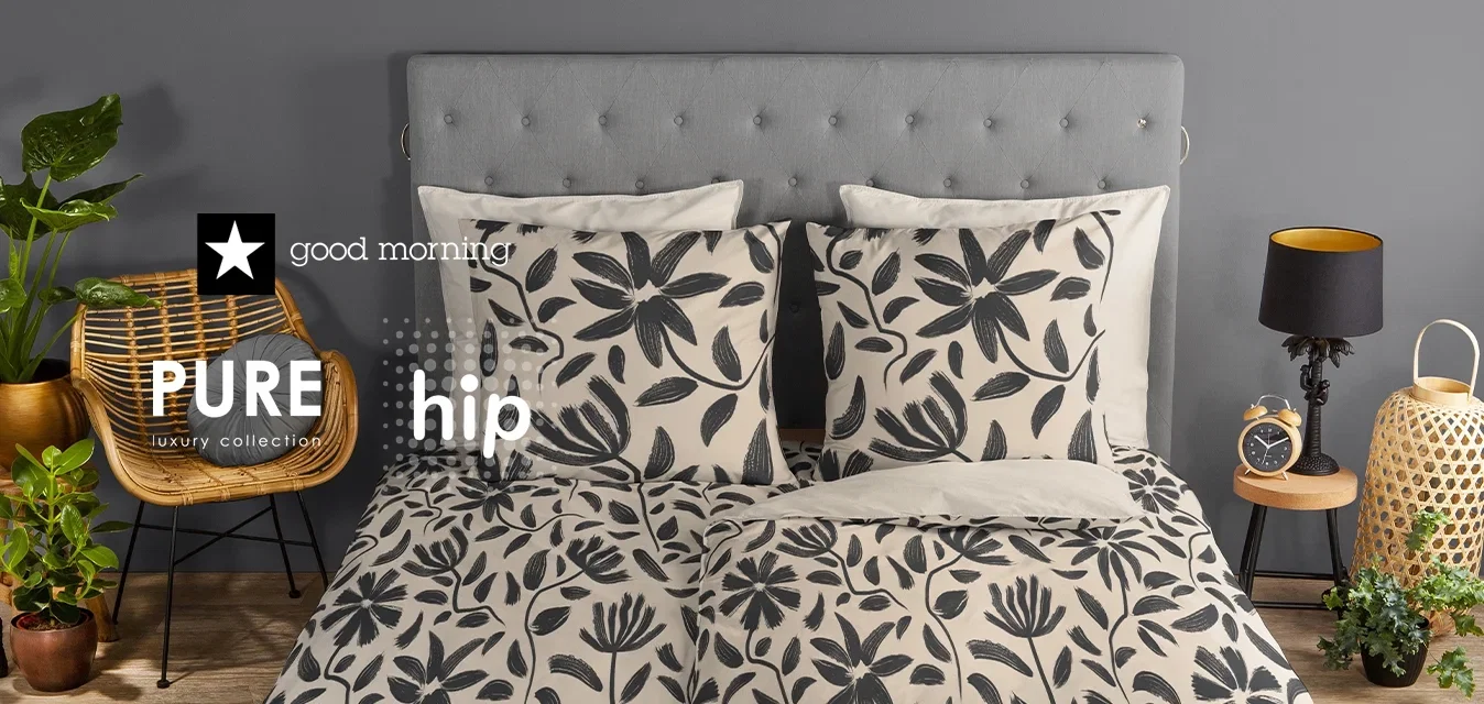 In einem Schlafzimmer steht ein grau gepolstertes Bett mit einem blumenmusterierten Bettbezug in Schwarz und Creme, kombiniert mit passenden Kissen. Geflochtener Stuhl, Pflanzen und eine Lampe.