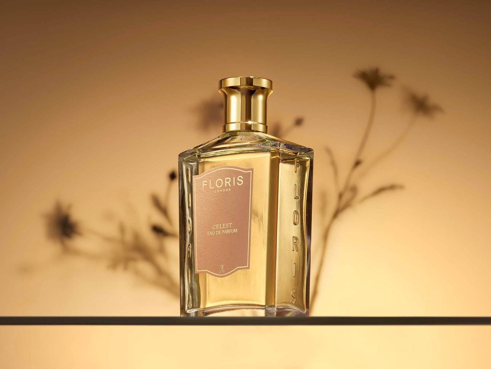 A bottle of Floris Celest Eau de Parfum with a gold cap is displayed on a glass shelf, against a softly lit background with blurred floral shadows.