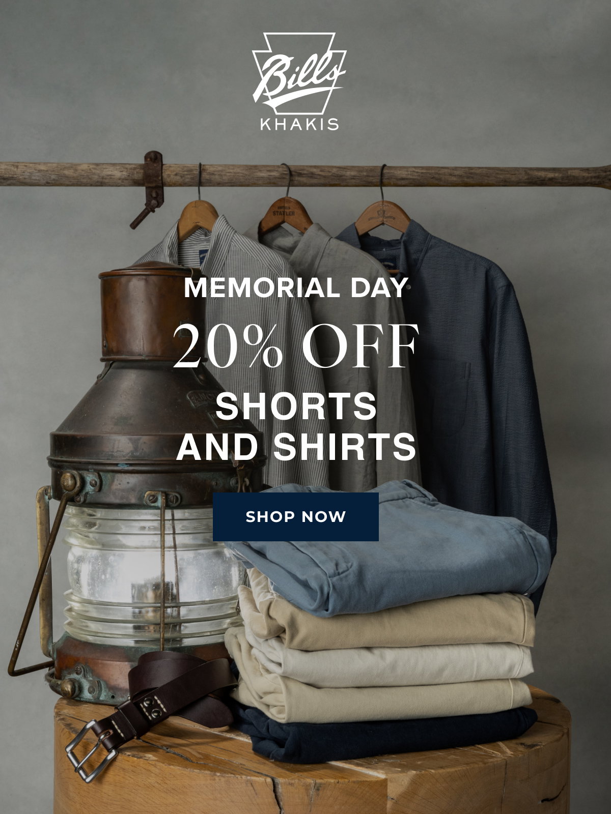 A background photo shows stacks of Bills Khakis shorts alongside hanging Bills Khakis shirts alongside vintage nautical objects . The Bills Khakis logo overlays the photo with text that reads: Memorial Day | 20% Off | Shorts and Shirts | Shop Now