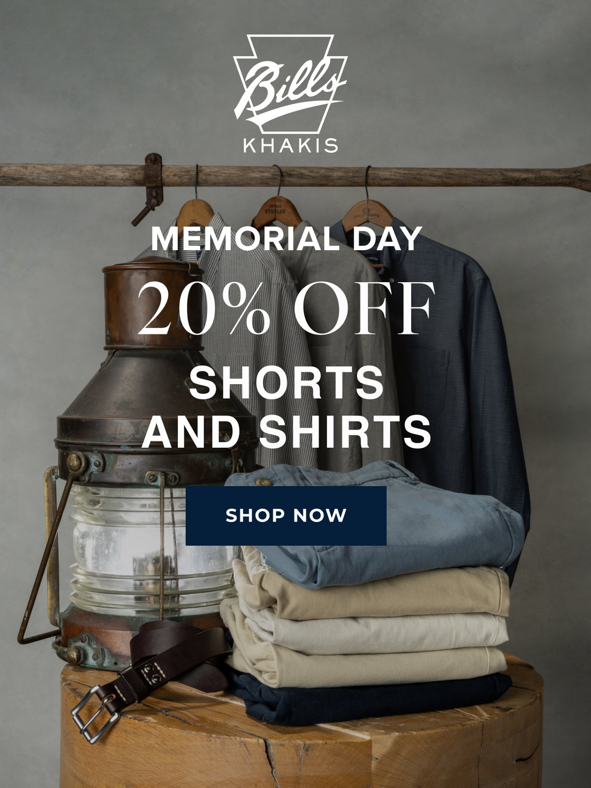 A background photo shows stacks of Bills Khakis shorts alongside hanging Bills Khakis shirts alongside vintage nautical objects . The Bills Khakis logo overlays the photo with text that reads: Memorial Day | 20% Off | Shorts and Shirts | Shop Now
