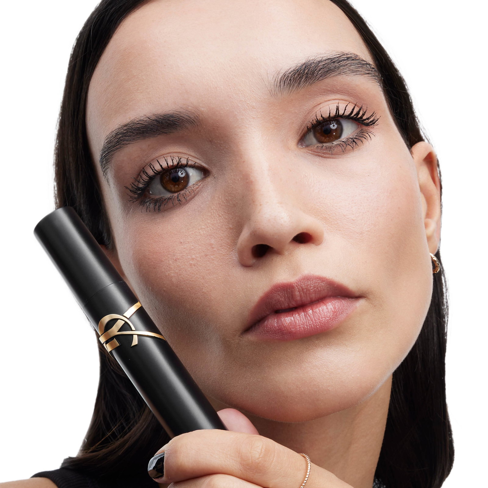 Close-up of a brunette model with bold, defined lashes holding the YSL Beauty Lash Clash mascara in its iconic black and gold packaging against a white background