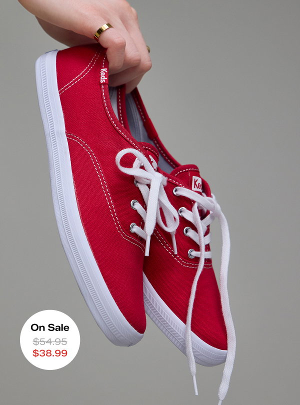 A grey background and a woman holding the Keds Champion Canvas Lace-Up in red. The style was $54.95 and is on sale for $38.99 for a limited time.