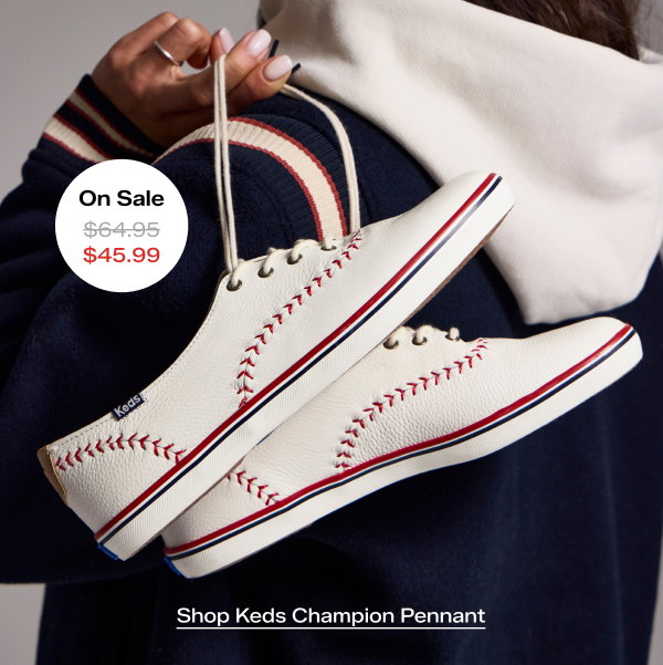 A woman is wearing a white hoodie and a navy wool bomber jacket with striped ribbing and is holding the Keds Champion Pennant Leather Lace-Up in off white by the laces. The style was $64.95 and is on sale for $45.99 for a limited time. Shop Keds Champion Pennant.