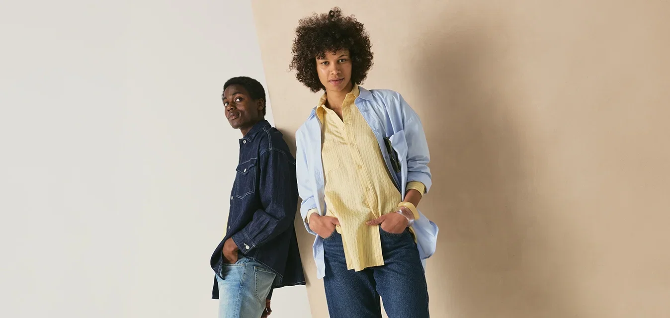 Due persone stanno appoggiate in modo informale a un muro; una indossa una camicia di jeans blu e jeans, l'altra una camicia azzurro chiaro, una maglietta gialla e jeans.
