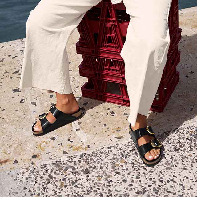 Person wearing white pants and black sandals sits on stacked red crates by the water.