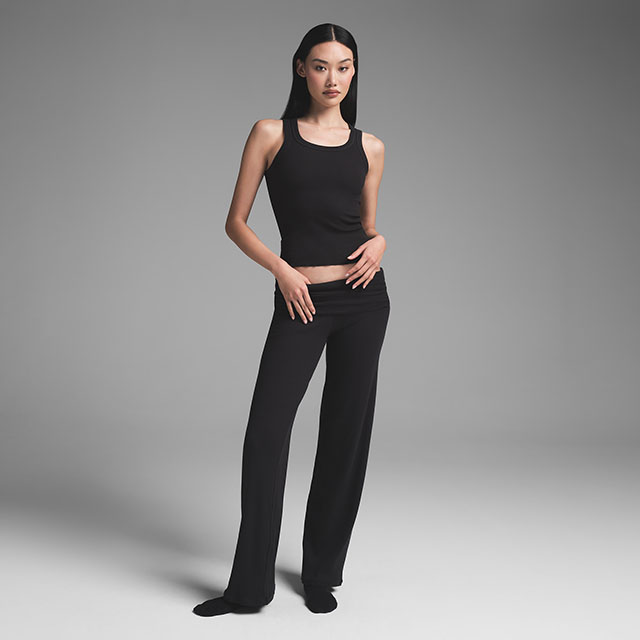 A person in a black sleeveless top and pants poses against a gray background.
