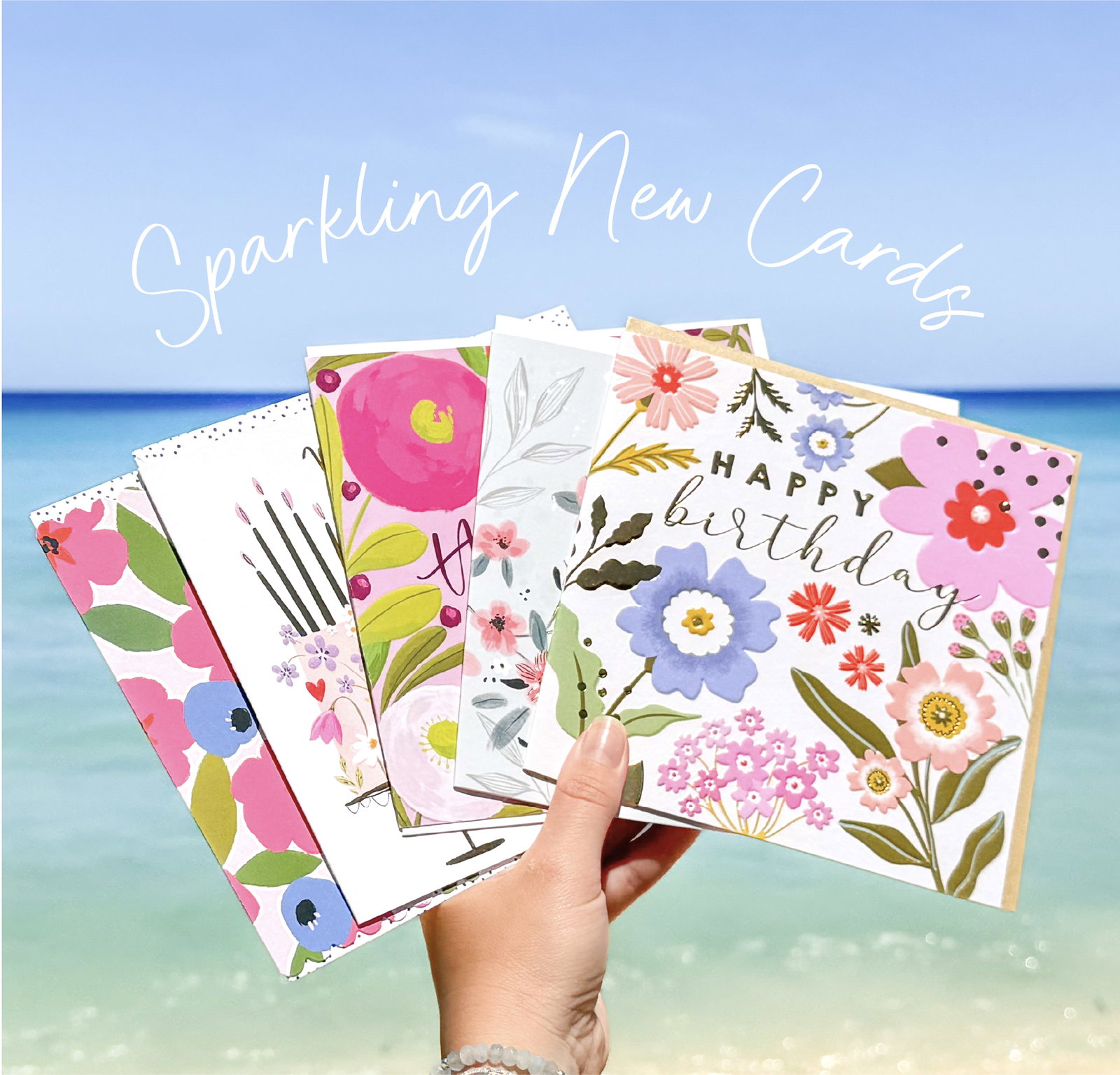 Hand holding a fan of bright floral and birthday greeting cards against a sparkling blue seaside background with handwritten text celebrating sparkling new cards for the season.
