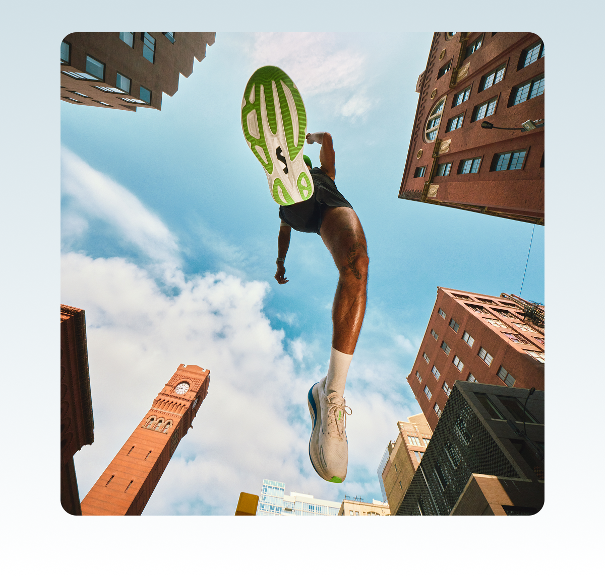 Low-angle image of a man jumping. The sole of his shoe is visible at the centre of the image, with buildings on either side and a blue sky in the background.