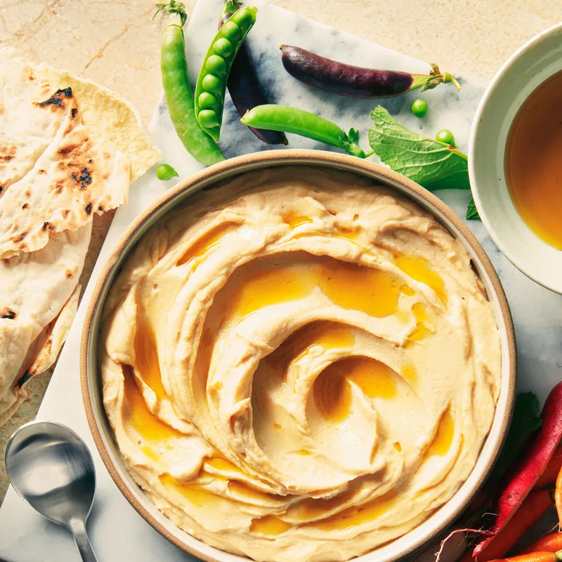 A bowl of hummus drizzled with olive oil surrounded by pita and vegetables.