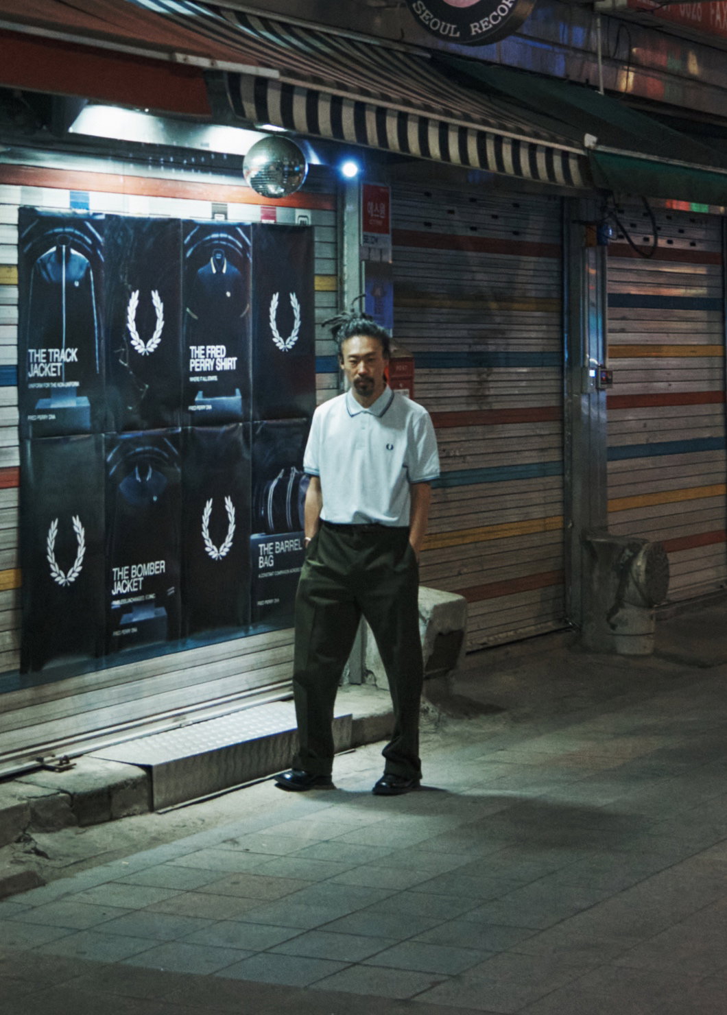 A Korean man stands in the distance on a dimly lit street in Soeul in front of Fred Perry posters and wears the Fred Perry Shirt in White / Light Ice / Navy. His long hair is tied back and his hands are tucked into the pockets of his black tailored trousers.