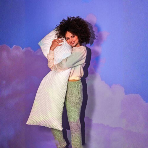 A smiling woman with curly hair hugging a long, white body pillow against a blue and purple background with cloud imagery.