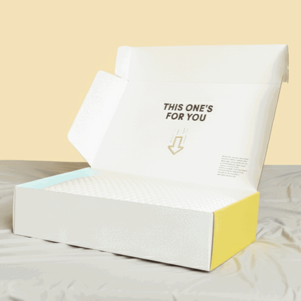 A white pillow inside a partially yellow and white open shipping box with the text 'THIS ONE'S FOR YOU' printed on the inner lid.