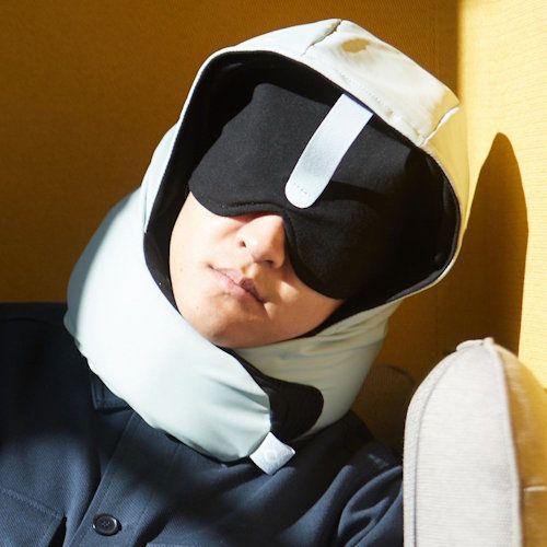 A person wearing a black sleep mask and a light blue travel neck pillow with a hood, resting in a yellow chair.