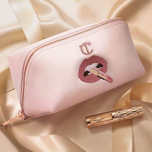A pink makeup bag with a zipper, featuring an embroidery of lips holding a lipstick. A gold lipstick tube lies next to the bag. The background is a soft, silky, light gold fabric.
