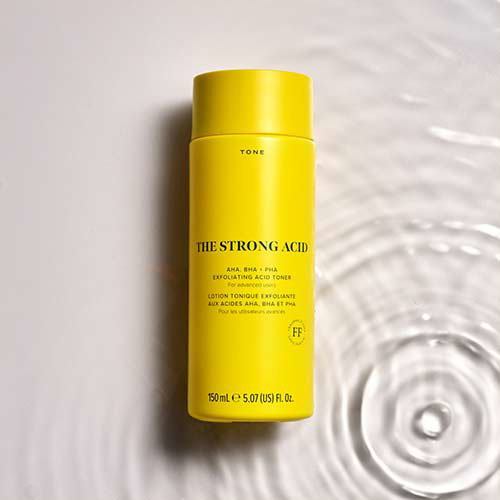 Bright yellow bottle of 'The Strong Acid' exfoliating toner with AHA, BHA, and PHA, placed on a rippled water surface reflecting soft light. The bottle has text detailing its features and contains 150 ml (5.07 fl oz).