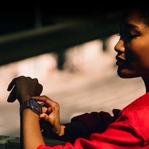 A person wearing a red jacket checks the display on a smartwatch, seated outdoors in a shaded area. Their attention is focused on the watch, with blurred surroundings in the background.