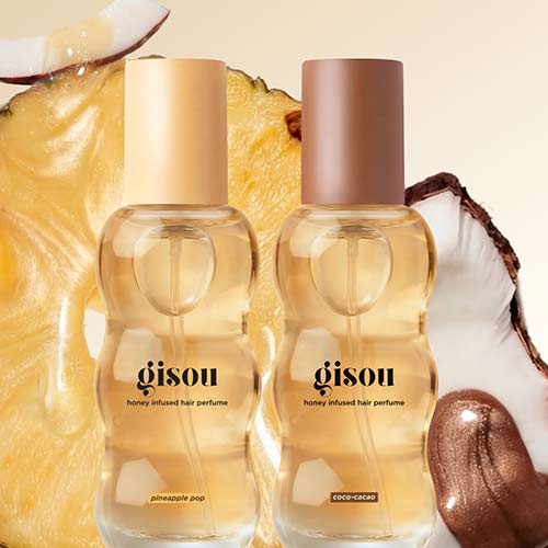 Two bottles of Gisou honey-infused hair perfume are displayed against a background featuring sliced pineapple and coconut. One bottle has a golden cap labelled 'Pineapple Pop,' and the other has a bronze cap labelled 'Coco-Cacao.' 