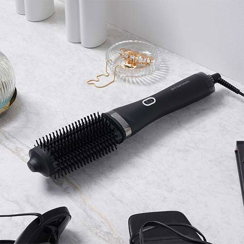 A black rotating hot air brush rests on a white marble surface. Nearby are a clear hair clip, a pair of sunglasses, and some bottles. The brush is plugged in, ready for use.
