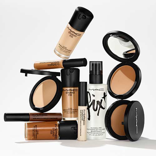 A selection of MAC makeup products including foundation bottles, concealer tubes, a setting spray, and compact powders with mirrors, arranged aesthetically against a white background.