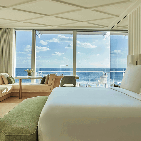 Hotel bedroom with a seating area by large windows, and a balcony overlooking the ocean.