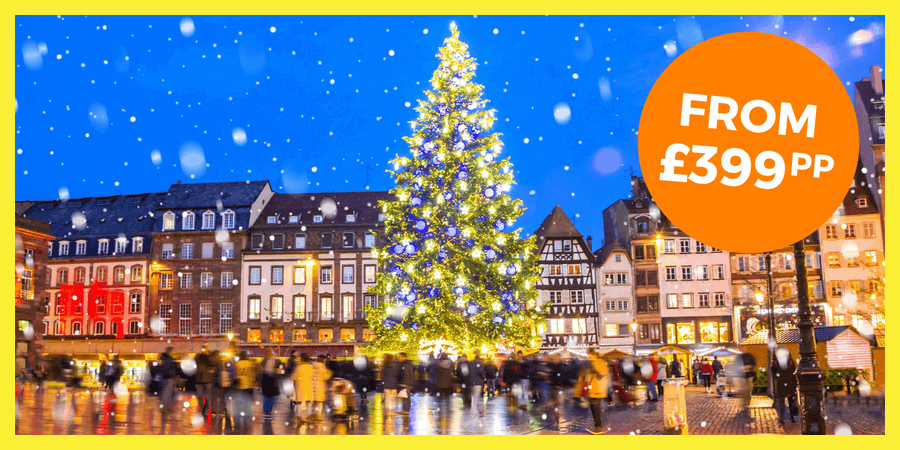 A festive Christmas tree adorned with lights stands in a bustling square, with snow gently falling.
