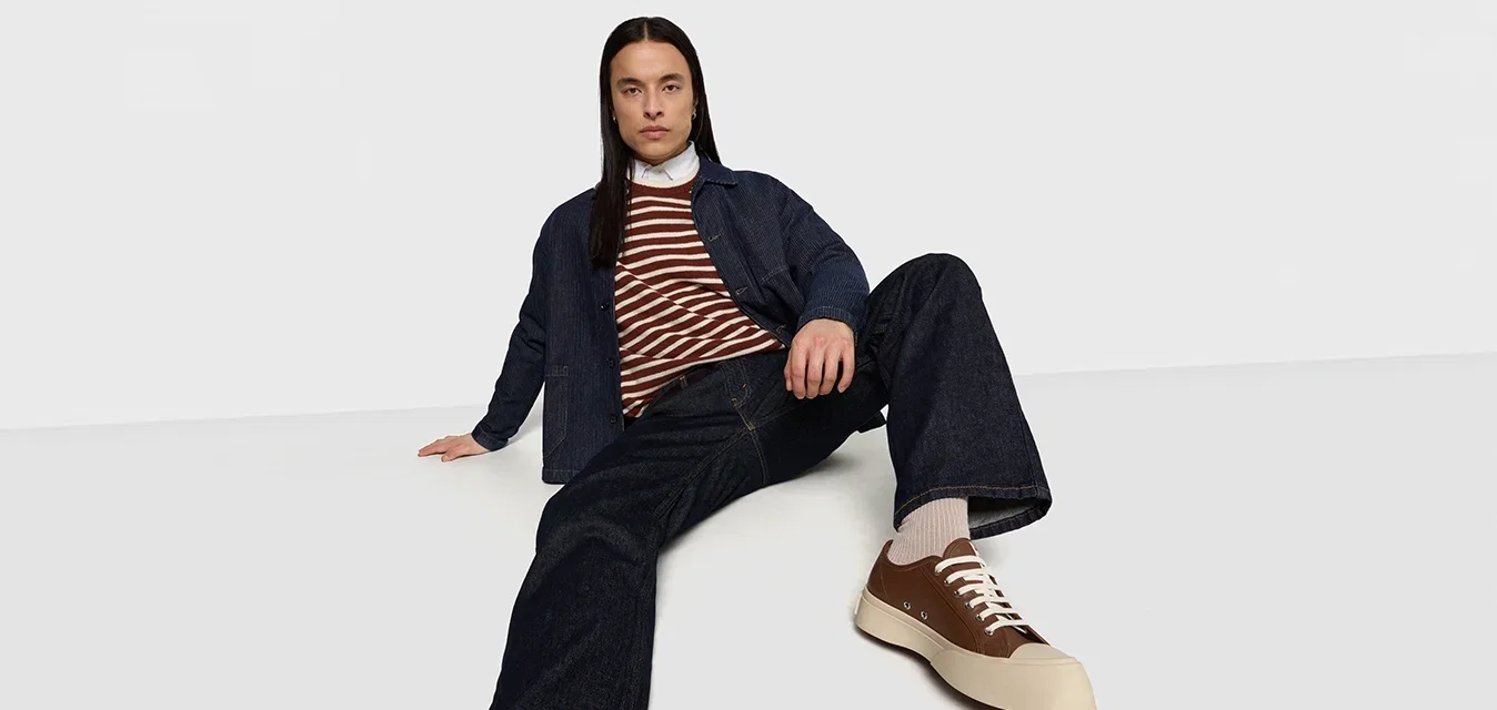 Chaqueta de mezclilla, suéter a rayas marrón y blanco, jeans oscuros, zapatillas beige con detalles marrones, en pose sentada, fondo neutro.