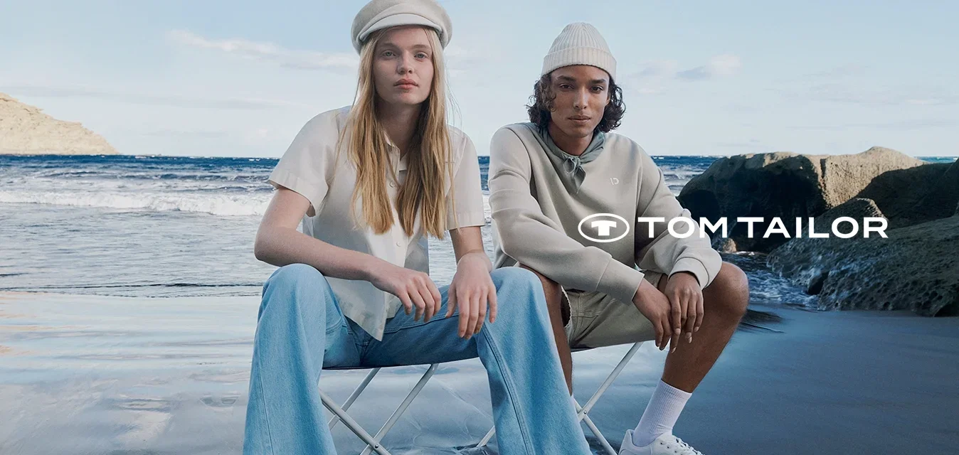 Twee personen zitten op opvouwbare stoelen bij het strand. De ene draagt een wit shirt en lichte jeans; de andere draagt een beige hoodie en korte broek. Beiden hebben witte sneakers aan.