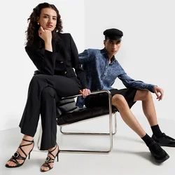 Zwei Models posieren in einem minimalistischen Setting. Eine trägt einen schwarzen Anzug und schwarze Riemchen-Heels; die andere trägt ein blaues Hemd und schwarze Shorts und sitzt.