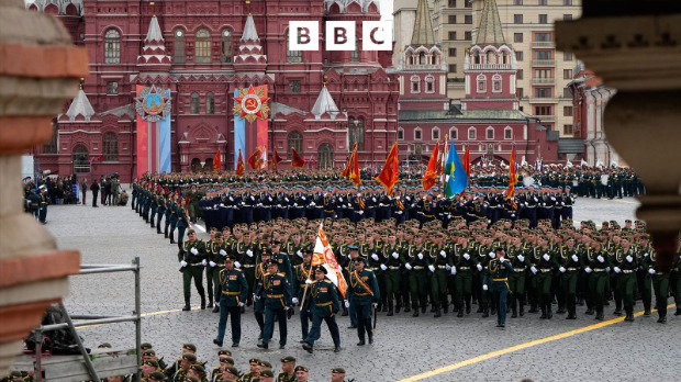 An image of a military parade in Moscow’s Red Square. Hundreds of military officers stand in formation, with some holding flags. There is a large red building in the background. There is a white BBC logo at the top of the image.
