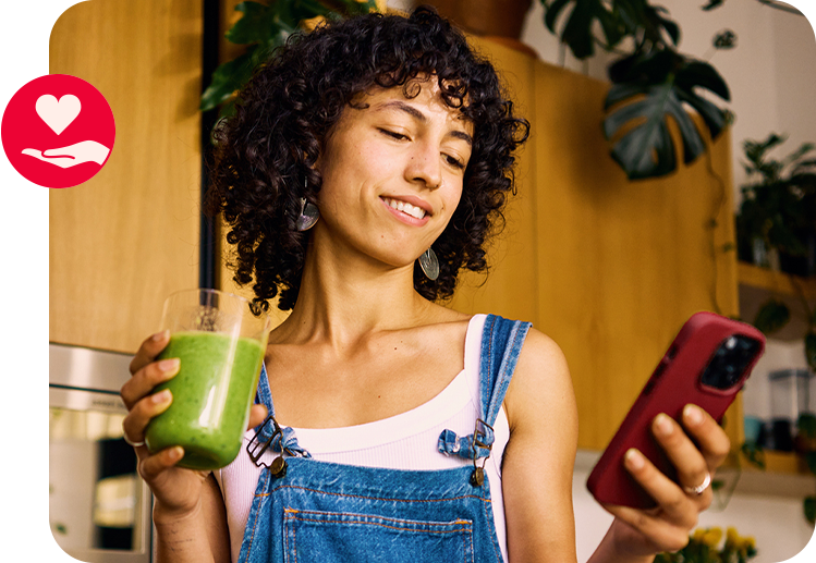 Frau hält grünen Smoothie und Smartphone in der Küche – Symbol für gesunden Lifestyle und Wohlbefinden.