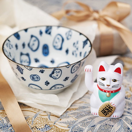Ceramic white bowl with blue lucky cat pattern next to a lucky cat ornament, with gift and ribbon in the background. - Shop Homeware.