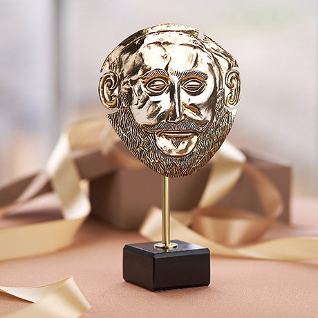 Gold-plated Funerary Mask of Agamemnon replica on a marble base with bands of gold ribbon in the background. - Shop Replicas.